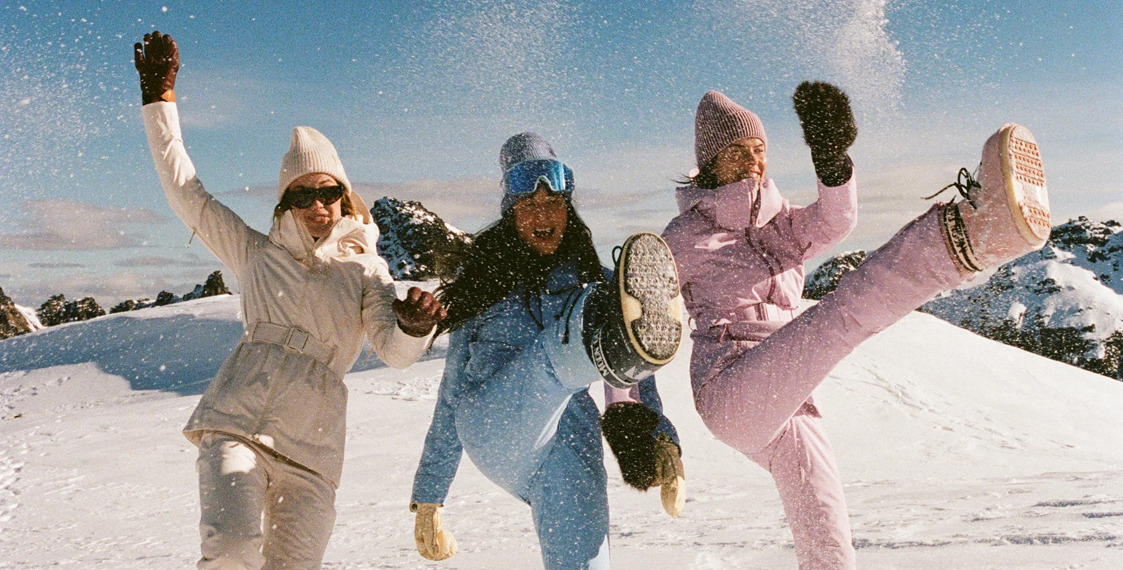 Three women in snow gear kicking up their legs on a snowy slope